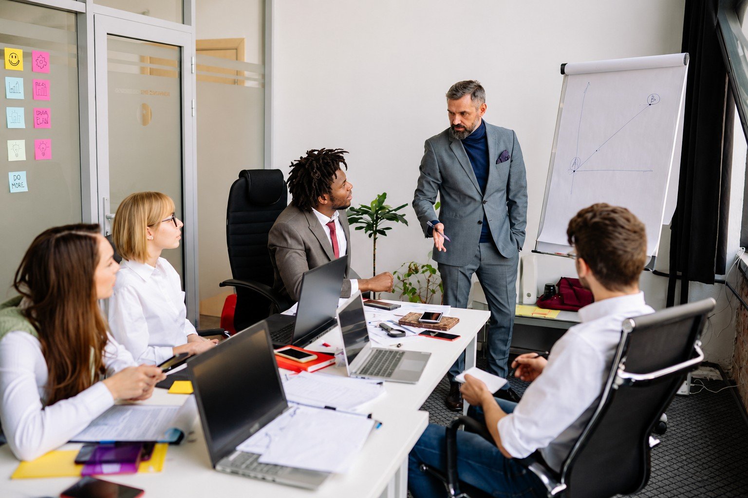 A business team discussing plans around a meeting table, with a colleague presenting ideas on a flip chart.
